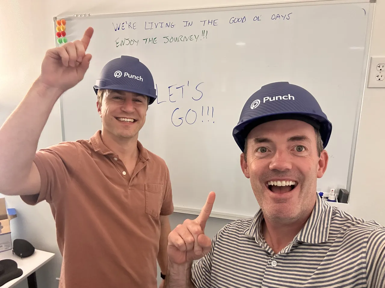 Taylor and Bradley grinning in Punch hard hats in front of a whiteboard that reads "We're living in the good ol' days — enjoy the journey!"